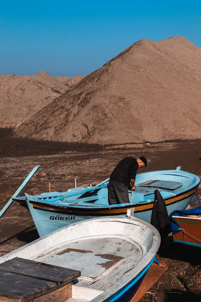 services-02 A man repairs a blue boat on a sandy beach under a clear blue sky, with mounds of sand in the background.